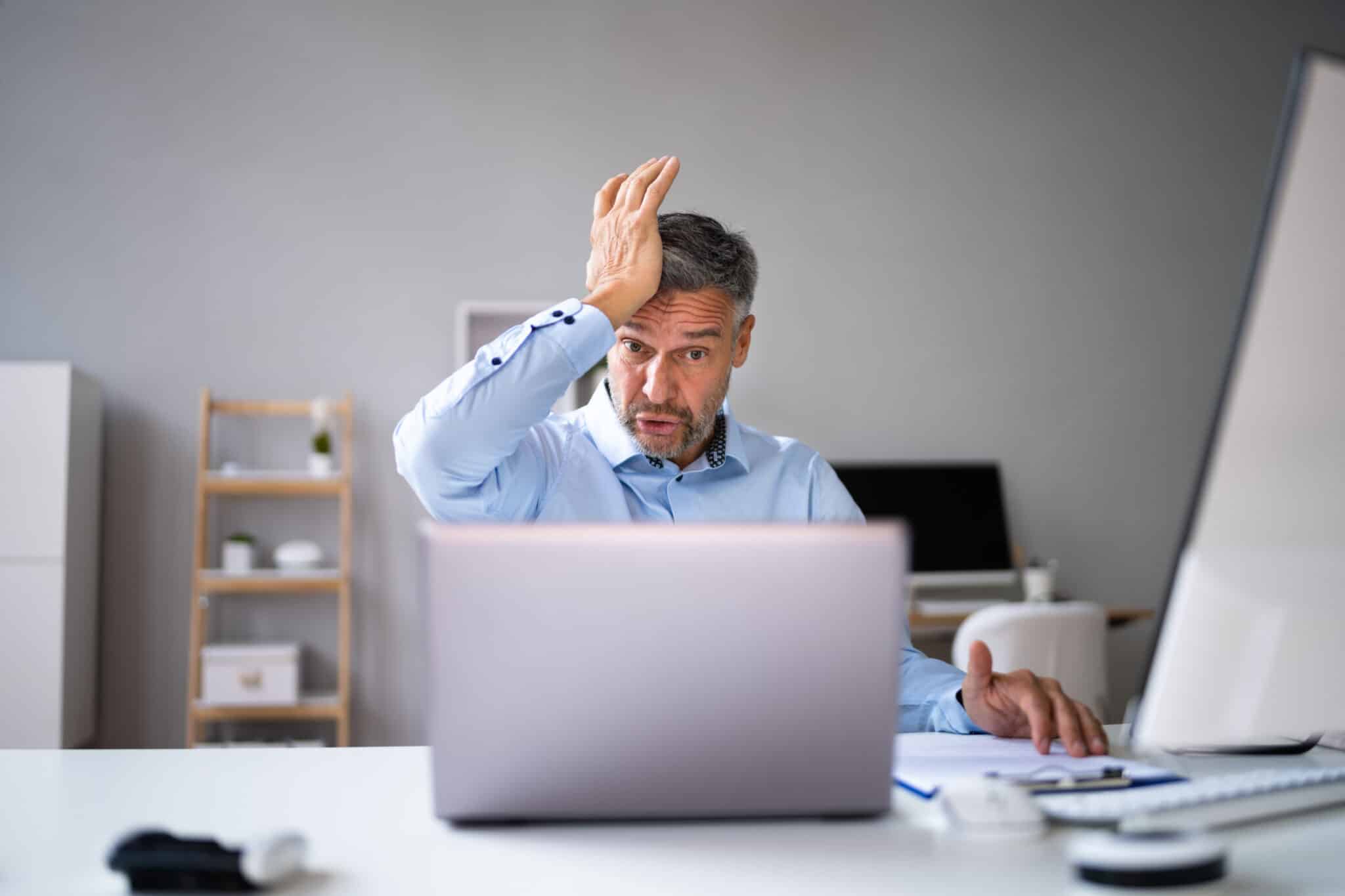 Concerned employee at desk with his hand on his head, looks at laptop screen, faces malware attack.