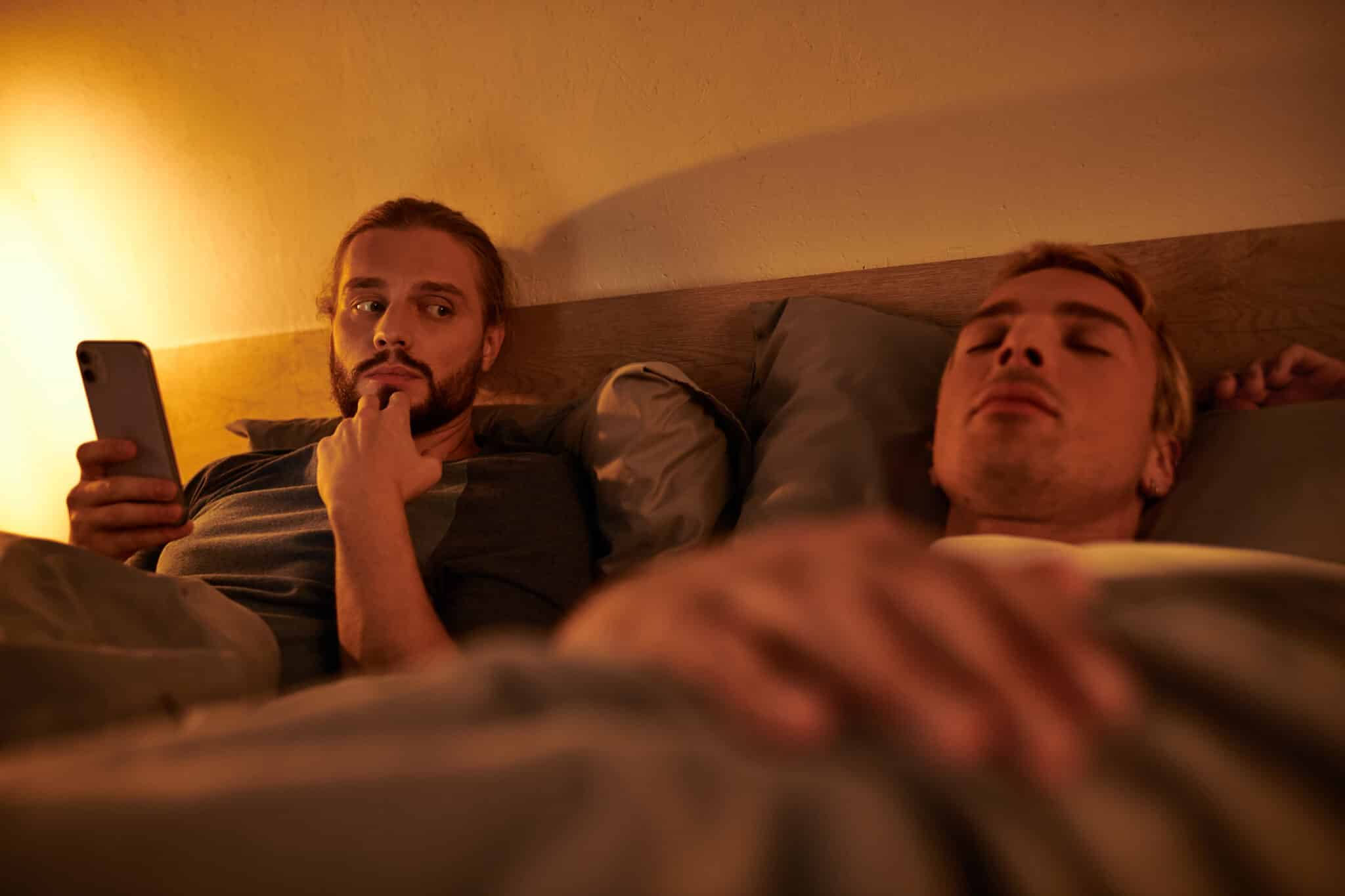 Man in bed secretly uses phone as partner sleeps; symbolizes secrecy & Ashley Madison breach lessons