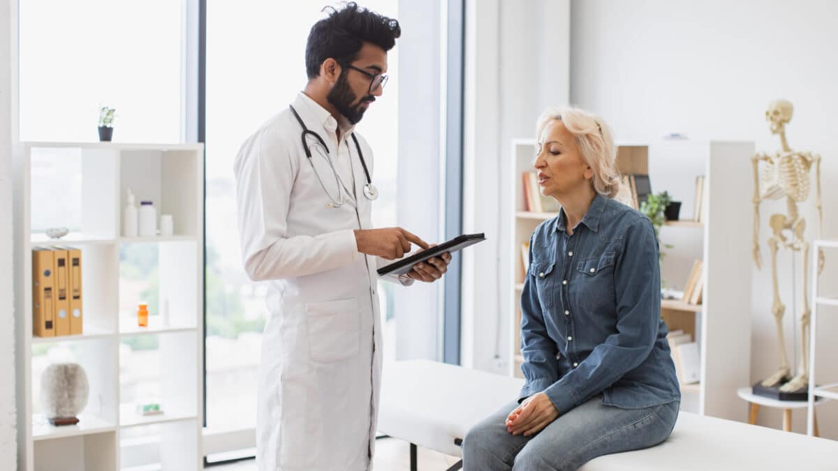 Doctor explains his use of an AI scribe to elderly patient while using his tablet in the exam room.