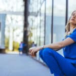 Tired doctor in scrubs sits outside hospital, symbolizes how AI scribes can reduce clinician fatigue