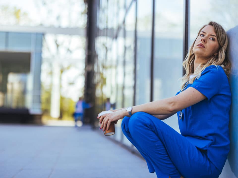 Tired doctor in scrubs sits outside hospital, symbolizes how AI scribes can reduce clinician fatigue