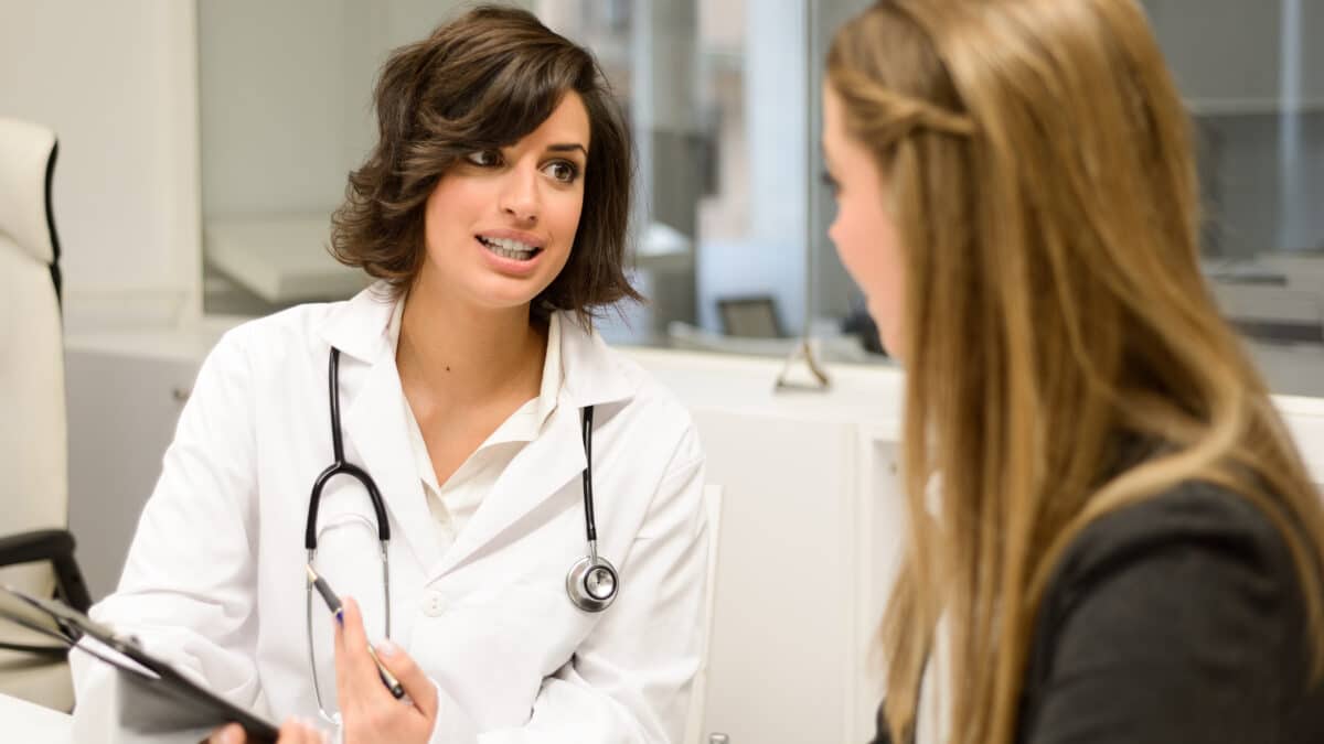 Doctor in white coat speaks with a patient, holding a clipboard, in a modern medical office setting.