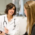 Doctor in white coat speaks with a patient, holding a clipboard, in a modern medical office setting.