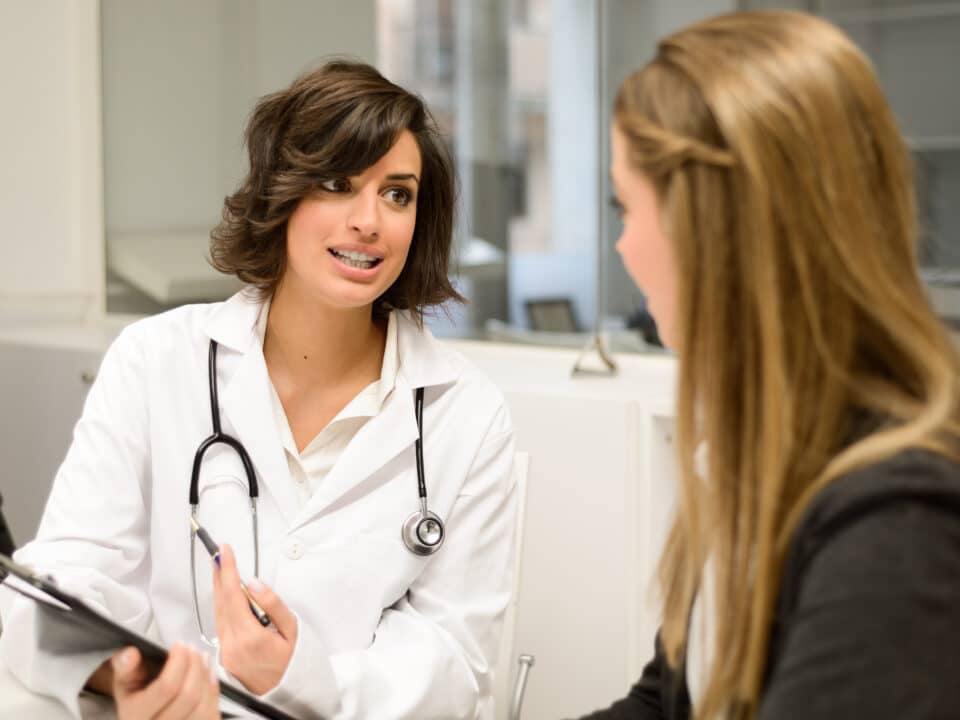 Doctor in white coat speaks with a patient, holding a clipboard, in a modern medical office setting.